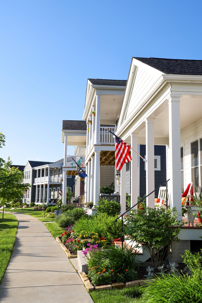Row of well-kept homes featuring front porches, gardens, and American flags along a sunny suburban sidewalk – JP Buys SD