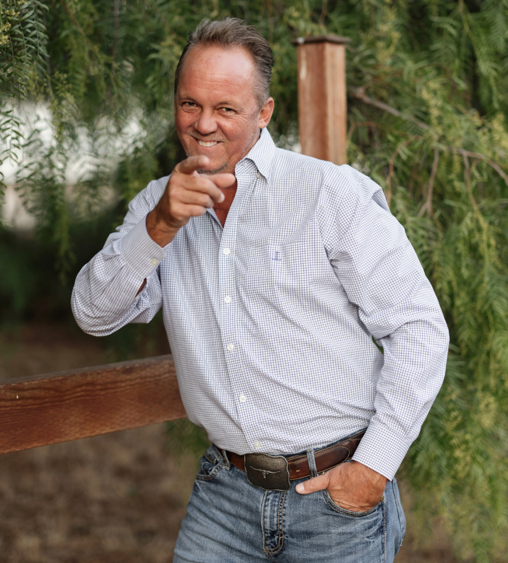 Jarrett Pugh in a button-down shirt and jeans pointing toward the camera while standing near a wooden fence and greenery – JP Buys SD