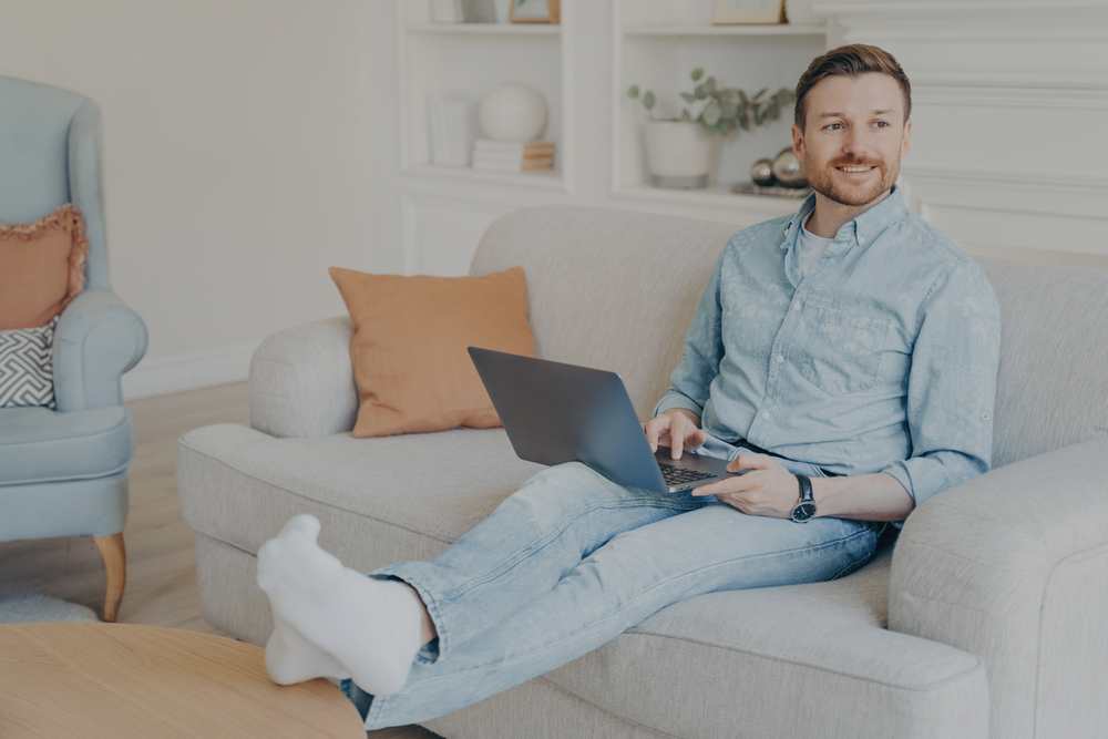 Smiling man sitting comfortably on a sofa with a laptop on his lap in a bright living room – JP Buys SD