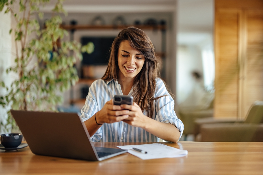 Smiling woman sitting at a desk using her phone beside a laptop and paperwork, representing professional communication and reliable property appraisal services – we buy houses vista